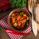 A bowl of stew featuring beef, tomatoes, yellow peppers, and fresh herbs sits atop a red checkered cloth, enriched with the robust flavor of The Spice Lab Organic Spanish Sweet Paprika. Surrounding the dish are chili peppers, parsley sprigs, bread slices, and wooden utensils arranged on a rustic wooden table.