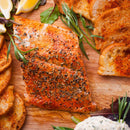 Seasoned baked salmon fillet on a wooden board, accompanied by toasted bread slices, lemon wedges, mixed greens garnished with The Spice Lab Fancy Chervil from The Spice Lab, and a small serving of creamy French-inspired dip.
