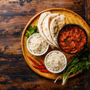 A woven tray holds naan bread, a bowl of curry seasoned with The Spice Lab Organic Ground Cumin, two bowls of white rice, red chili peppers, and fresh cilantro, all arranged on a rustic wooden table.