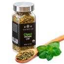 A glass jar from The Spice Lab Organic Starter Spice Set - 6 Jars sits next to a wooden spoon filled with dried oregano and a few fresh oregano leaves on a white background, making it an ideal addition to your everyday cooking spices or the perfect choice for any starter spice set from The Spice Lab.