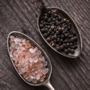 Two antique spoons rest on a rustic wooden surface, one filled with The Spice Lab Himalayan Pink Salt and the other brimming with The Spice Lab Premium Black Pepper from their Grinder Set.