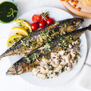 A plate features two grilled fish accompanied by a side of diced potatoes with herbs, cherry tomatoes, and lemon wedges. The potatoes are garnished with The Spice Lab Organic Parsley Flakes from The Spice Lab for added freshness. A green sauce and bread are also visible nearby.