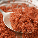 A close-up of a silver spoon holding The Spice Lab Hawaiian Red Alaea Sea Salt Medium Grain crystals, renowned for their distinctive earthy flavor, with more of this premium salt from The Spice Lab scattered in the background.