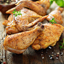 Succulent, roasted chicken pieces—drumsticks and wings—resting on a wooden table, garnished with fresh herbs and a bold dash of The Spice Lab Birdseye Chili Pepper Powder, complemented by scattered coarse salt and peppercorns.