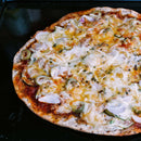 A baked pizza topped with melted cheese, green olives, red onions, and a sprinkle of The Spice Lab Ground Oregano sits on a black baking tray.