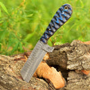 A Texan Knives Cowboy Bull Cutter with a 4" handmade Damascus steel blade and a blue-black textured handle is embedded in a log, lush green foliage in the background.