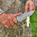 Wearing camouflage, a person uses the Texan Knives Custom Handmade Damascus Steel Skinner Knife with Sheep Horn Handle – 4.5" Blade to peel a plant stem, with blurred green foliage in the background.