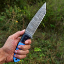 A hand holds the Texan Knives Custom Handmade Damascus Steel Skinning Knife with a 4.5" blade and resin handle outdoors, against blurred green foliage.