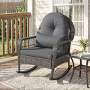 The Patio Rattan Rocking Chair with Cushions - 400 LBS Capacity by DirectDeals adds style to your porch, featuring gray cushions next to a glass table with flowers, atop a patterned rug with plants in the background.