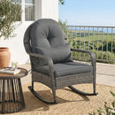 The DirectDeals Patio Rattan Rocking Chair with Cushions - 400 LBS Capacity sits on a patio beside a side table with a book and drink, surrounded by greenery, potted plant, rug, railing, and clear blue sky.