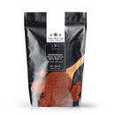 A black bag labeled The Spice Lab Hawaiian Red Alaea Sea Salt Fine Grain rests beside a wooden spoon overflowing with vibrant red salt, with some crystals scattered on the white surface. This 1 lb package from The Spice Lab is ideal for anyone looking for premium fine grain sea salt to enhance their culinary creations.