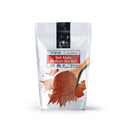 A white package labeled The Spice Lab Hawaiian Red Alaea Sea Salt Medium Grain showcases an image of red salt with a distinctive earthy flavor spilling from a wooden spoon, with the brand name The Spice Lab displayed at the top.