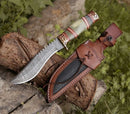 The Texan Knives Handmade bowie knife features a hand forged Damascus steel blade and a multicolored handle, shown resting on a log with its detailed brown leather sheath—an ideal collectible for custom knife enthusiasts.