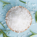 A wooden bowl filled with The Spice Lab Spanish Sea Salt Coarse Grain sits on a light blue surface, surrounded by scattered salt crystals and sprigs of fresh rosemary—an inviting scene enhanced by the distinctive texture of this premium sea salt from The Spice Lab 3-Pack with Grinder.