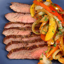 Sliced grilled steak, seasoned with The Spice Lab Grilling Seasoning Collection from The Spice Lab, served with sautéed onions and red and yellow bell peppers, garnished with chopped herbs on a dark plate.