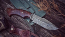 The Texan Knives Good Hunting Knife, featuring a 10" Damascus steel blade, rosewood handle, and leather sheath, rests on a green metal box outdoors among dry twigs and weathered boards.