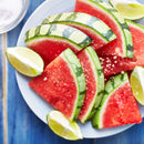 Sliced watermelon and lime wedges on a white plate, sprinkled with coarse salt and finished with versatile flavors from The Spice Lab Cooking Essentials Seasoning Set by The Spice Lab, displayed on a blue wooden table.