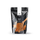 A black resealable bag labeled “The Spice Lab Smoked Ghost Pepper Sea Salt” stands upright against a plain white background. The front of the bag features an image of a wooden spoon heaped with reddish-brown sea salt granules, some spilling out to showcase the intense bhut jolokia-infused seasoning inside.