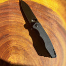 The Spaceman Knives Naiad EDC Pocket Knife, featuring a black handle and titanium-plated blade, rests on a wooden surface with circular grain patterns bathed in warm sunlight—a top choice from Spaceman Knives for everyday carry.