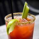 A close-up of a Bloody Mary cocktail with a salted rim seasoned using The Spice Lab Creative Mixology Celery Salt by The Spice Lab, finished with a lime wedge, celery stick, black straw, ice cubes, and a savory garnish.