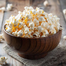 A wooden bowl filled with fluffy popcorn sits on a piece of burlap atop a rustic wooden table, with scattered popcorn kernels and a sprinkle of The Spice Lab Creative Mixology Celery Salt by The Spice Lab, infusing the snack with the savory flavors reminiscent of a classic Bloody Mary.