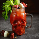 A Bloody Mary served in a skull-shaped glass with a cocktail rim seasoned using The Spice Lab Creative Mixology Celery Salt, and garnished with celery, olives, pickles, salami, and eggs on skewers—all arranged on a dark surface.
