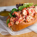 A lobster roll with mixed greens in a toasted bun sits on parchment paper, accompanied by a small metal cup of melted butter and a sprinkle of The Spice Lab Creative Mixology Celery Salt from The Spice Lab, evoking the classic flavor of cocktail rim seasoning.