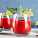 Two glasses of red Bloody Mary cocktails with salted, seasoned rims—each made with The Spice Lab Creative Mixology Celery Salt by The Spice Lab—are garnished with celery stalks and lime wedges, sitting on a light surface against a blue background. Nearby, a bowl of green olives and slices of lime complement the classic touch provided by The Spice Lab’s celery salt in each Bloody Mary.