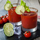 Two glasses of Bloody Mary cocktails garnished with lime slices and crisp celery, their rims dusted with The Spice Lab Creative Mixology Celery Salt. Cherry tomatoes, lime slices, and ice cubes are scattered across the black slate surface nearby.