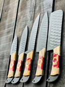 Five Texan Knives Stainless Steel Knife Set 13" with forged blades and olivewood & epoxy handles are arranged in a row on a rustic wooden surface, each accompanied by a leather sheath.