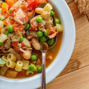 A close-up of a bowl of vegetable soup featuring pasta, white beans, peas, tomatoes, and herbs, infused with The Spice Lab Organic Fennel Seeds for a subtle licorice flavor, with a spoon resting in the bowl on a wooden table.