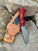 The Texan Knives Hunting Skinning Knife features a 10" Damascus steel blade and pakkawood handle, shown with its leather sheath resting on large gray rocks outdoors.