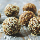 Six round energy balls coated with The Spice Lab Organic Hulled Sesame Seeds and oats rest on a rustic wooden surface. Shredded coconut is scattered around, creating a natural and homemade snack vibe.