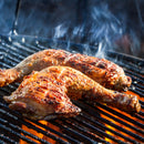 Two pieces of grilled chicken with crispy, browned skin are cooking on a hot barbecue grill over bright orange flames, with smoke rising around them. Infused with Caribbean-inspired flavor and finished with The Spice Lab Scotch Bonnet Sea Salt by The Spice Lab, these chicken pieces promise a bold and spicy kick in every bite.