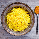 A decorative bowl filled with cooked yellow rice sits on a wooden surface next to a spoon holding The Spice Lab Organic Ground Turmeric, a bright yellow staple in Indian cooking.