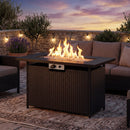 A 44.5″ Propane Fire Pit Table with 50,000 BTU Burner by DirectDeals glows with flames, surrounded by wicker patio furniture on an outdoor rug at sunset. String lights and plants accent the stone wall behind.