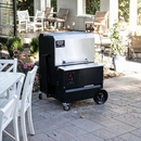 A Myron Mixon BARQ 2400 Pellet Smoker (BARQ-2400) with Wi Fi from Myron Mixon Smokers sits on a stone patio near a white table and chairs, surrounded by lush greenery and sunlight.