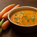 A bowl of creamy carrot soup garnished with fresh parsley and a sprinkle of The Spice Lab Whole Leaf Cilantro sits next to three whole carrots on a wooden surface.