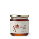A glass jar of Georgetown Olive Oil Co Raw Unfiltered Varietal Honey—an exceptional California varietal honey—with a gold lid and a label featuring a pink flower illustration. This raw and unfiltered honey is presented in a 9.5 oz (269g) net weight.