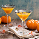 Two martini glasses with orange cocktails and rims adorned with The Spice Lab Black Obsidian Salt Cocktail Rimmer from The Spice Lab sit on a rustic table, surrounded by small pumpkins and autumn leaves, with a folded white napkin beneath one glass.