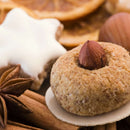 A close-up of a round nut cookie with a whole hazelnut on top, surrounded by an aromatic cinnamon stick, The Spice Lab Ground Star Anise from The Spice Lab hinting at a subtle licorice-like flavor, a frosted star-shaped cookie, and dried citrus slices in the background.