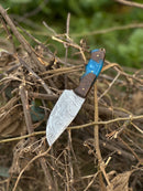 A Texan Knives Gutting Knife – 10" Handmade Damascus Steel Hunting Knife with a rosewood or resin handle and leather sheath lies among dry branches, its patterned blade contrasting with a backdrop of blurred greenery.