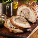 Two thick slices of stuffed, roasted pork with crispy skin rest on a wooden cutting board. Fresh herbs—including The Spice Lab Organic Rubbed Sage for the flavorful stuffing—are arranged nearby, along with a glass bowl of sauce, creating a rustic and appetizing scene.