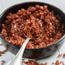 A close-up of a black bowl filled with The Spice Lab Hawaiian Red Alaea Sea Salt Medium Grain crystals, showcasing their medium grain, reddish-brown hue as a silver spoon scoops up some of the salt to highlight its distinctive earthy flavor.