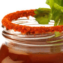 A close-up of a glass rimmed with The Spice Lab Bloody Mary Salt Cocktail Rimmer by The Spice Lab and chili powder, filled with a reddish savory cocktail and garnished with a celery stalk.