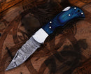 The Texan Knives Personalized Damascus Steel Pocket Knife features a blue and black handle with metal rivets, resting on carved wood—a perfect Custom Valentines Day Gift Knife for Husband.