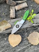 The Texan Knives Handmade Skinner Knife features an 8" Damascus steel blade and a pakkawood wire-cut handle, shown resting on a wooden log with dry and green leaves, paired with a leather sheath.