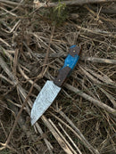 The Texan Knives 10" Handmade Damascus Steel Hunting Knife with a resin and wood handle rests on dry, tangled sticks and grass.