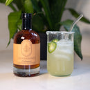 A bottle of Under The Bark Syrup Co Habanero Maple Syrup sits beside an iced, pale green jalapeño drink with a salted rim and glass straw, while a leafy plant blurs softly in the background, enhancing the sweet heat vibe.