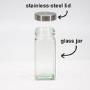 A square empty French glass spice jar from The Spice Lab 12 Pack Empty French Glass Spice Jars set sits below a hovering stainless-steel lid. Black arrows and labels identify both the durable lid and the elegant glass jar by The Spice Lab, highlighting their use for stylish seasoning storage against a clean white background.